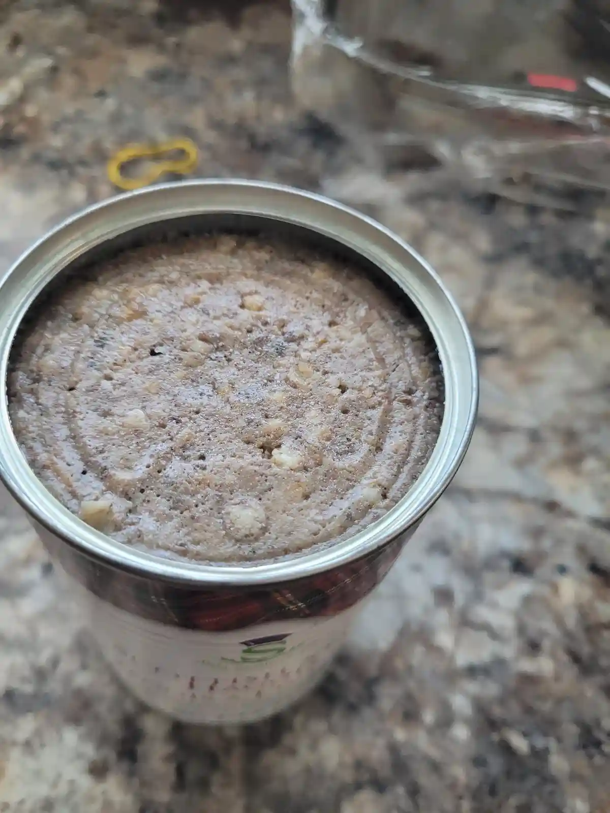 A top-down view of an open metal can sitting on a speckled countertop, revealing a brown, textured, cake-like substance…