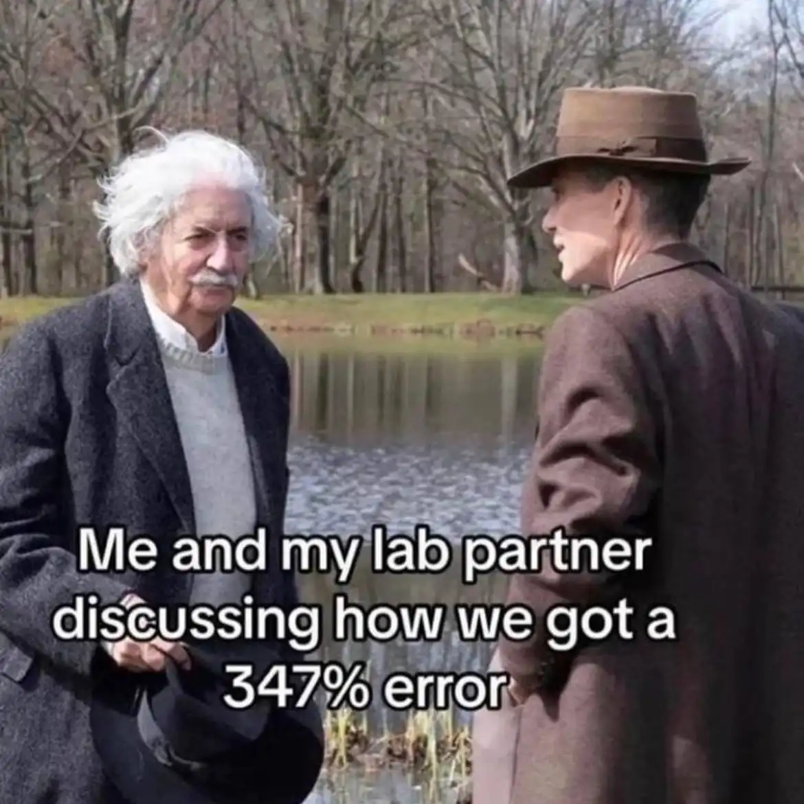Two men stand by a pond in a wooded area; one with wild white hair and a mustache, the other in a brown hat and coat. The…