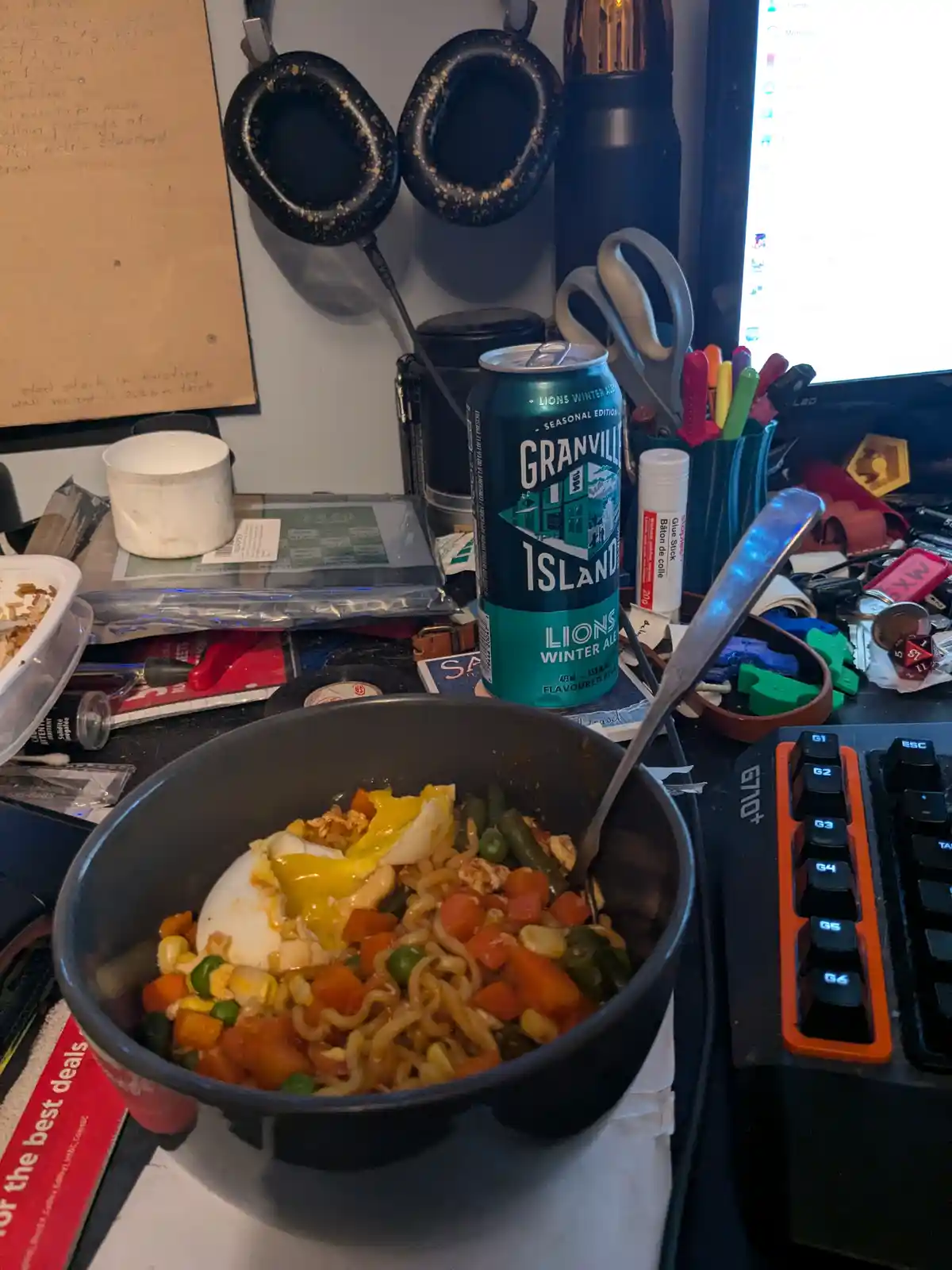 A cluttered desk features a black bowl of ramen with mixed vegetables and a cracked hard-boiled egg on top, next to a can of…