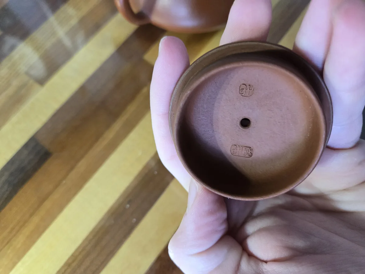 A hand holds a small, brown ceramic tea pot upside down, revealing its bottom with a central hole and two embossed maker's…
