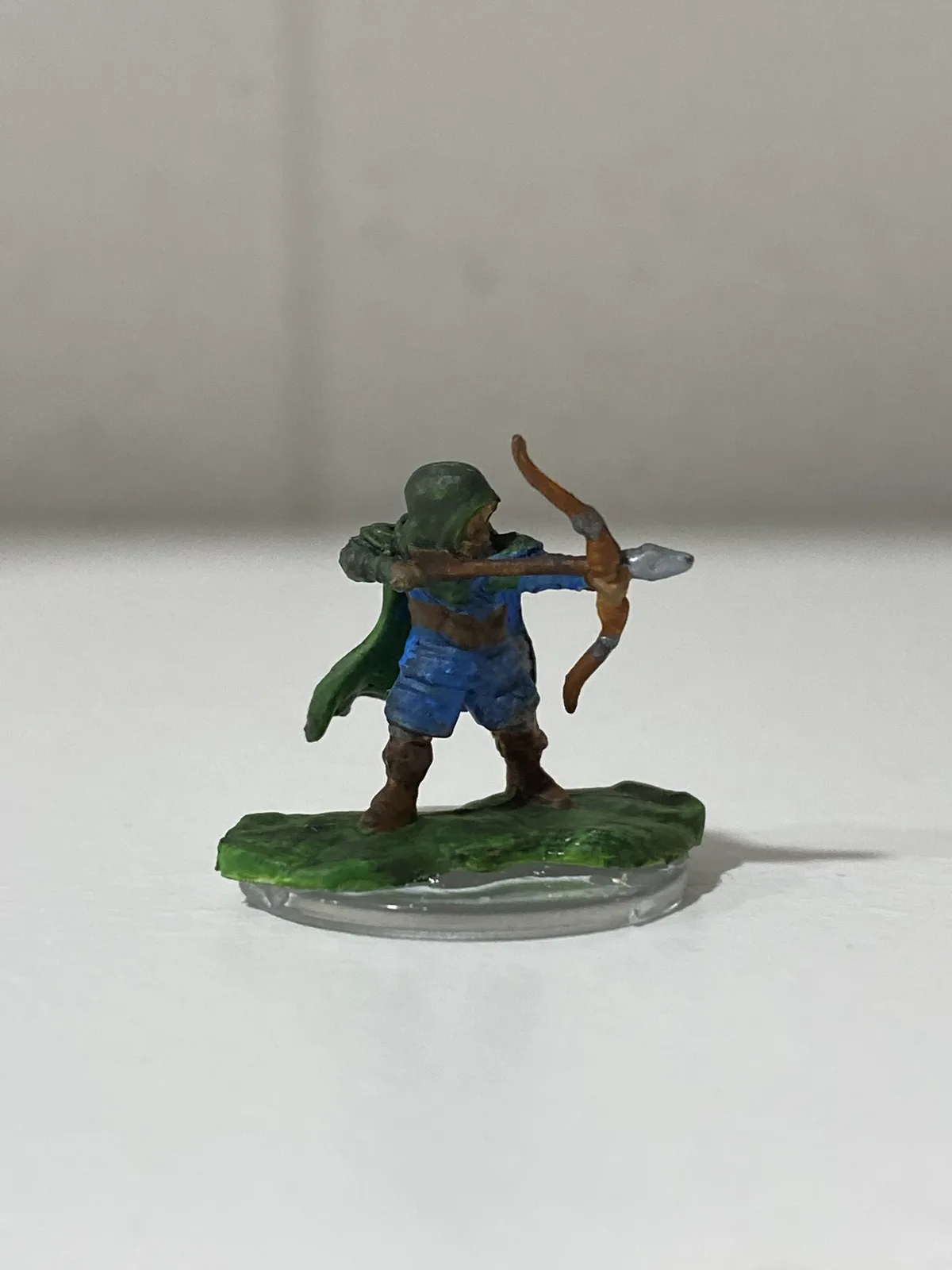 A painted miniature figurine of a hooded archer in blue and green attire, standing on a green base and drawing a bow, with…