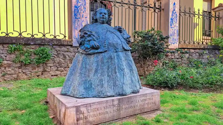 A blue-bronze statue of a very large, robed woman stands on a stone pedestal in a grassy area. Behind her is a stone wall…