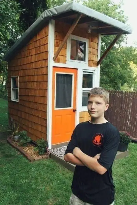 A young man with arms crossed stands proudly in front of a small, wooden, orange-door tiny house in a backyard. The house…