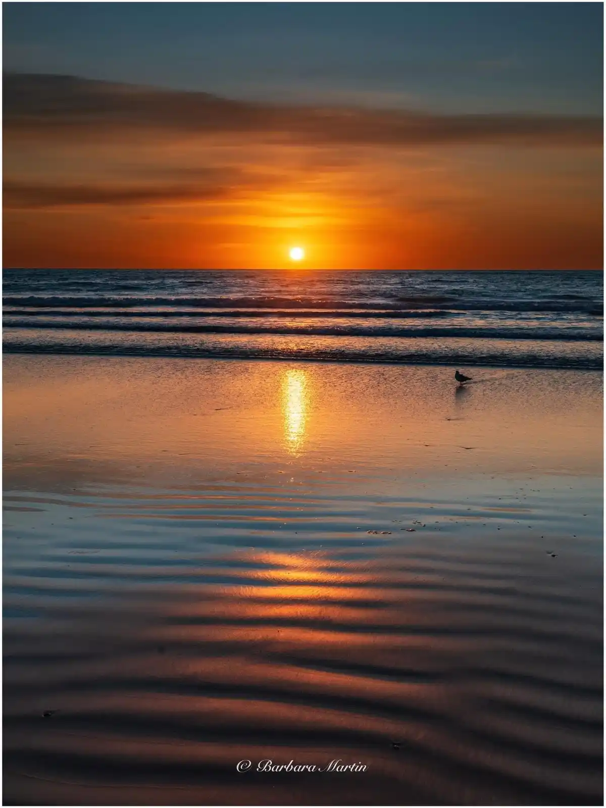 A serene beach sunset photo shows the sun setting over the ocean, casting a golden reflection on wet sand with gentle waves…