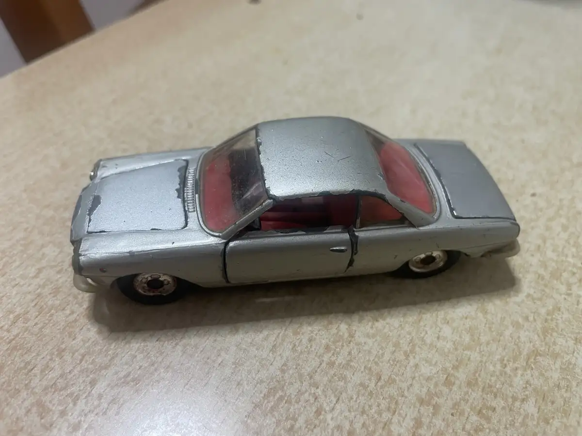 A silver toy car with visible damage, including a cracked roof and broken door, sits on a light-colored wooden surface. The…