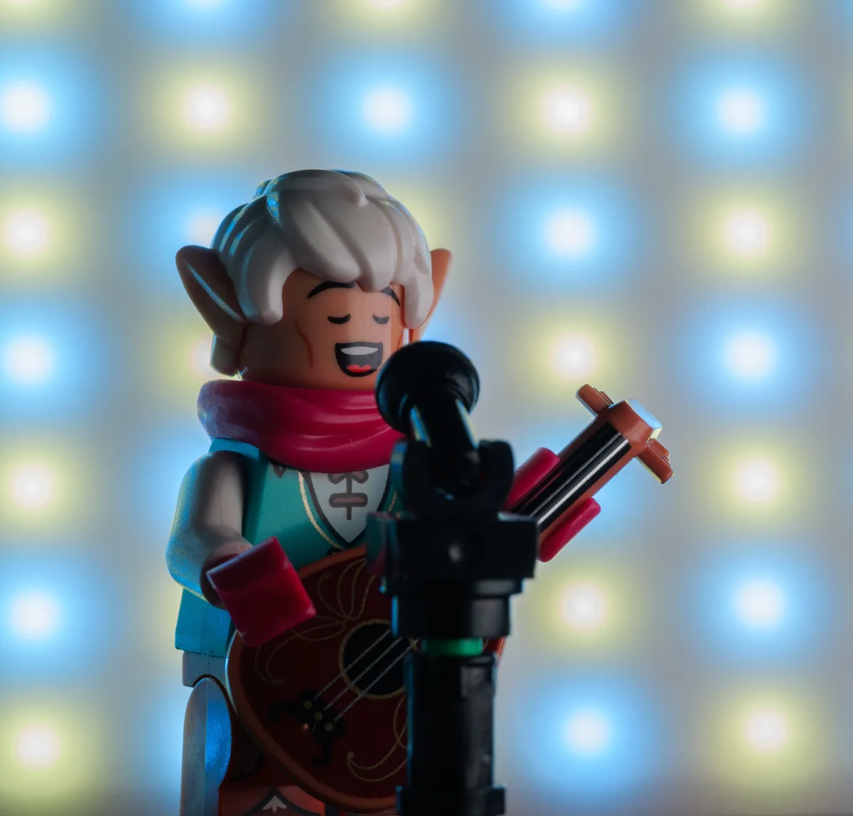 A Lego minifigure with elf ears, white hair, and a red scarf is posed as a singer holding a small guitar, standing before a…