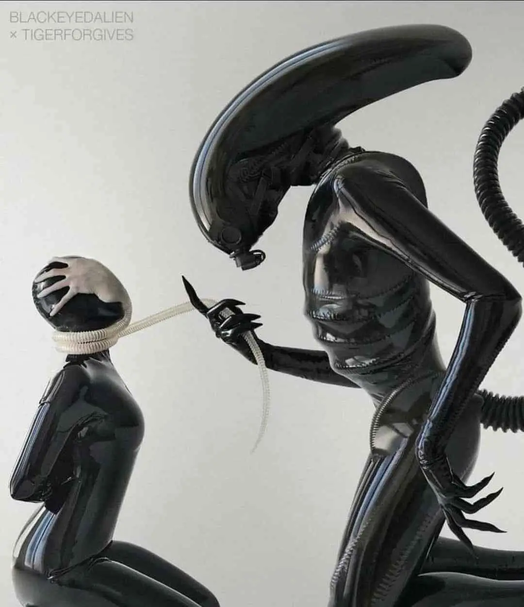 A glossy black alien figure with a long head crest and biomechanical features is shown kneeling and holding a white tube to…