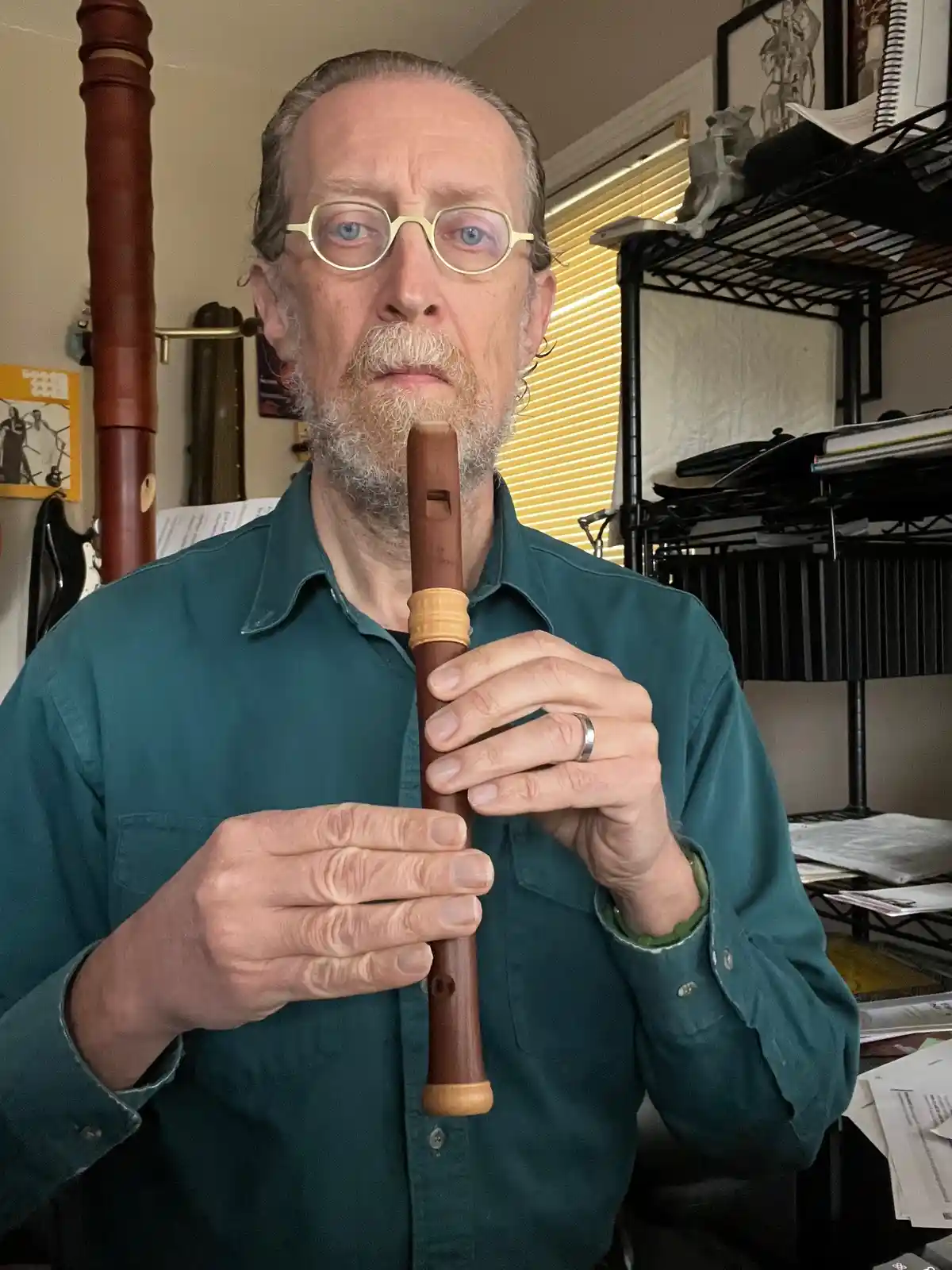 A middle-aged man with glasses and a beard holds a wooden flute to his lips, looking intensely at the camera. He wears a…
