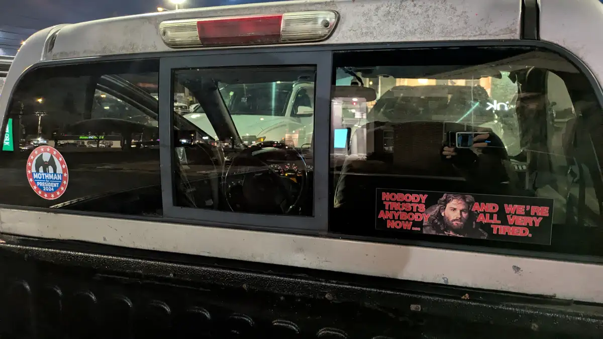 A nighttime photo of a white pickup truck's rear window, showing a 'Mothman for President 2024' sticker on the left and a…