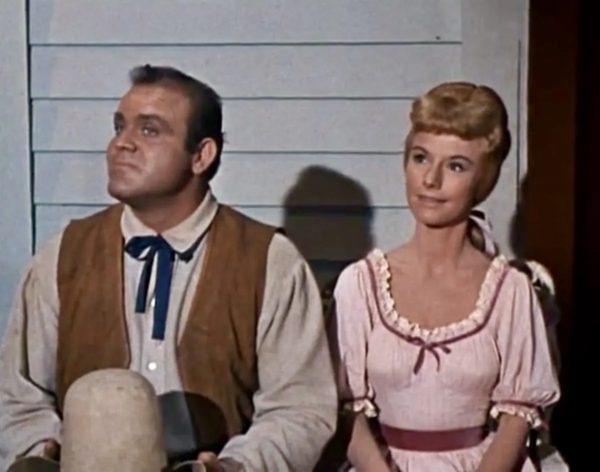 A man and a woman in vintage western-style clothing sit side by side, both looking upward with expressions of mild…