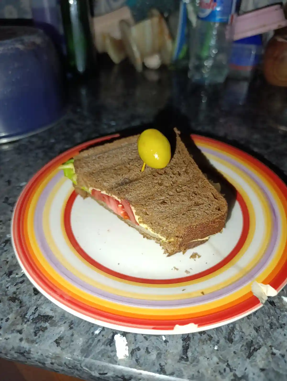 A triangular sandwich on whole wheat bread sits on a colorful striped plate, topped with a single green olive on a…