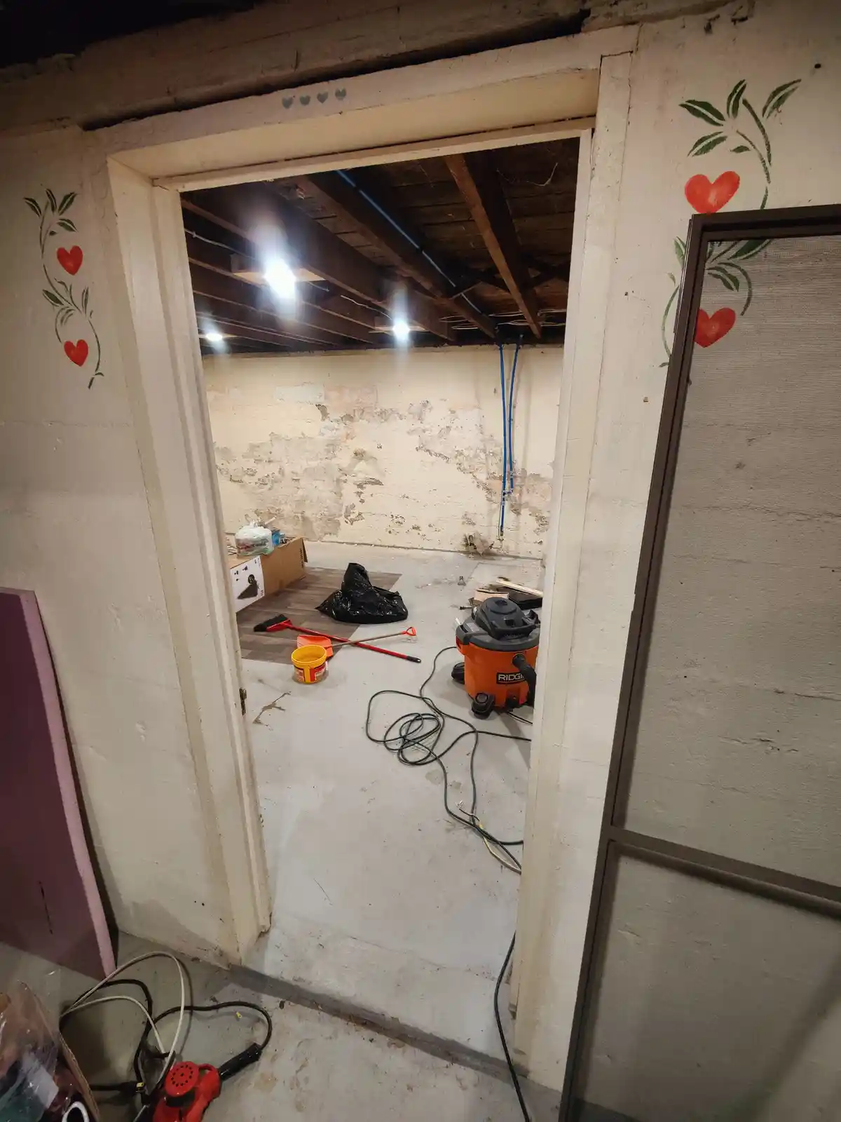 A doorway leads into a messy, unfinished basement with exposed beams, peeling walls, and scattered tools like a vacuum…