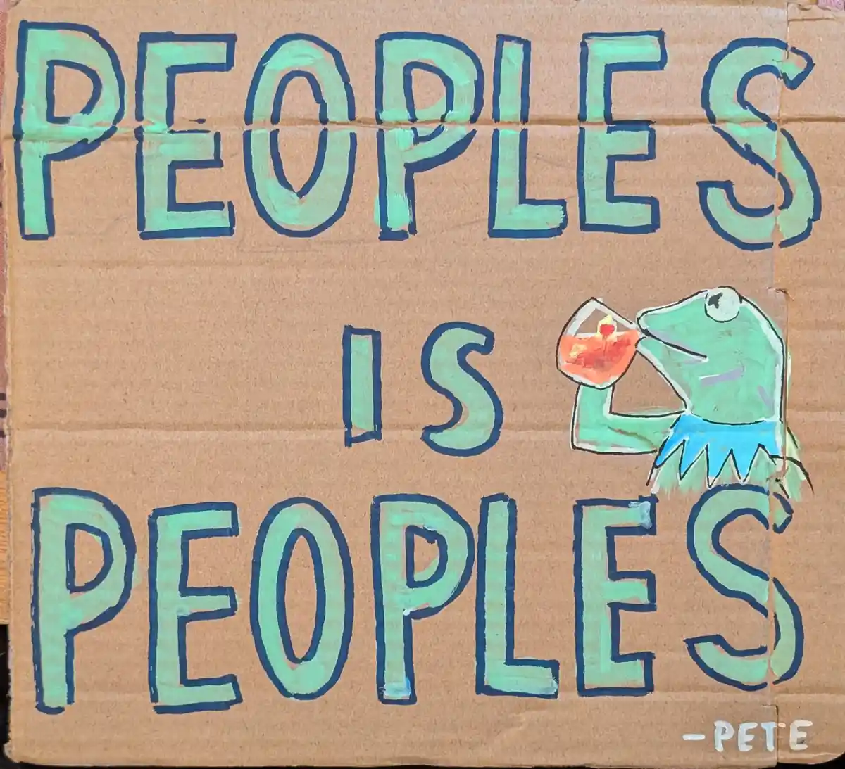 A hand-painted sign on brown cardboard displays the text 'PEOPLES IS PEOPLES' in large, teal letters with dark blue…