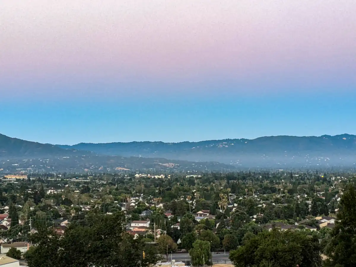 A scenic photo of a suburban landscape with houses, trees, and distant mountains under a gradient sky transitioning from…