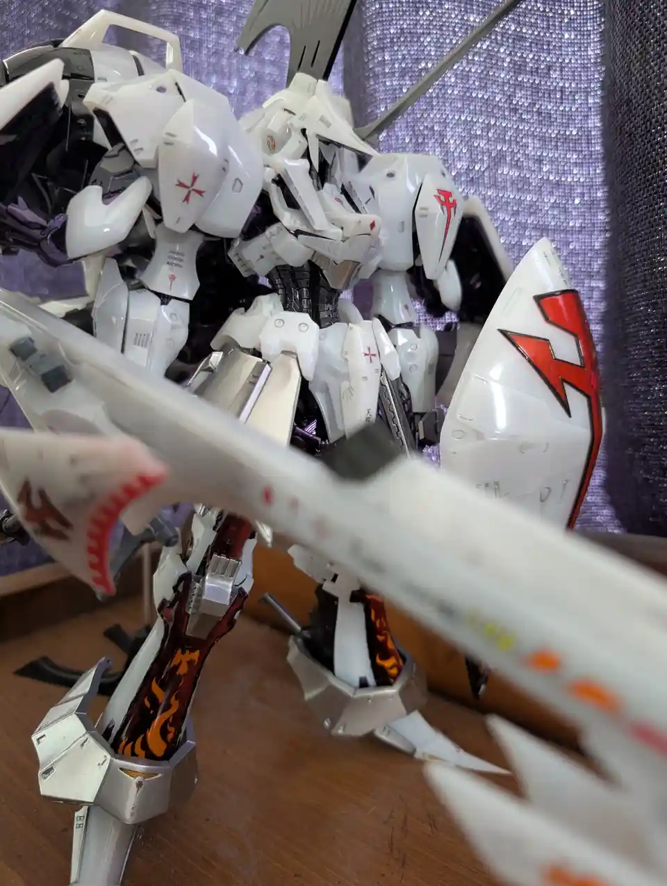 A close-up photo of a white and black mecha model with red markings, standing on a wooden surface against a purple textured…