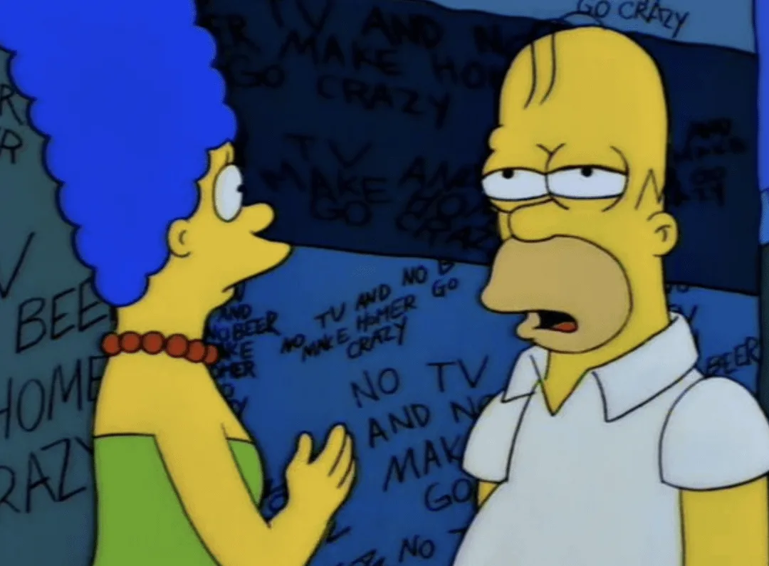 In this still from The Simpsons, Marge Simpson stands beside Homer, who looks annoyed, as she points to a wall covered in…