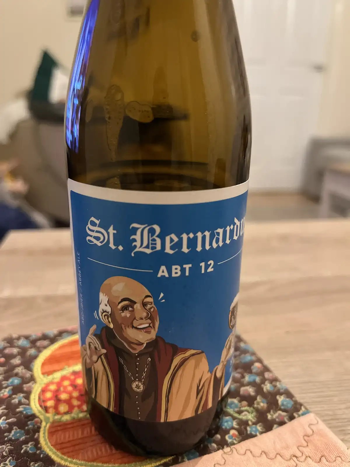 A close-up photo of a brown glass beer bottle with a blue label featuring a cartoon illustration of a smiling bald monk. The…
