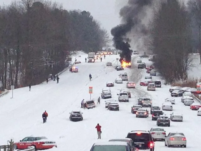 a snowy road is filled with cars and people, with a large fire and thick black smoke rising in the distance, and the text…