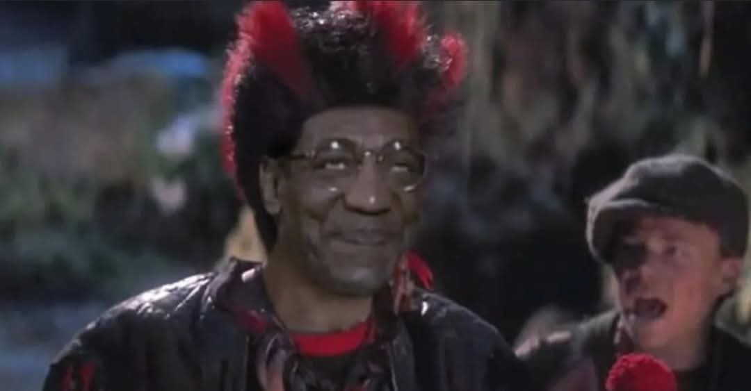 A man with a red and black mohawk wig and glasses smiles smugly while wearing a dark jacket over a red shirt. Behind him, a…