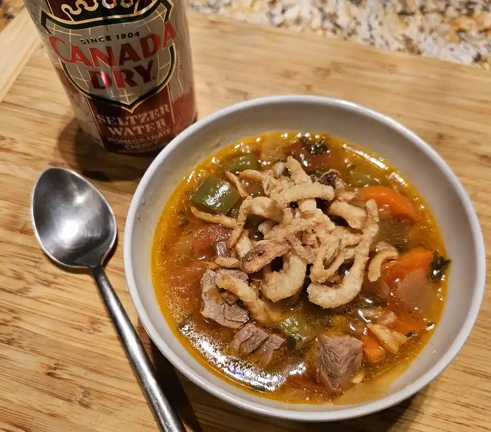 A bowl of hearty beef vegetable soup topped with crispy fried onions sits on a wooden surface next to a spoon and a can of…