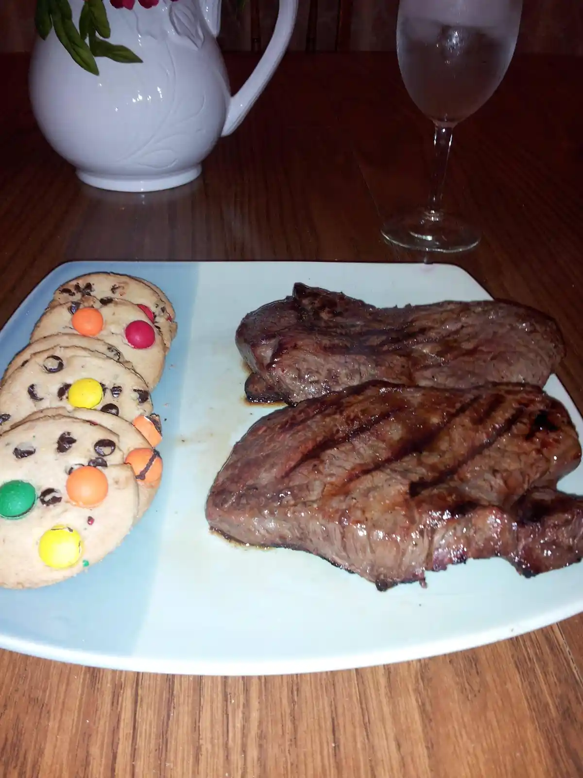 A plate of grilled steak sits next to a row of chocolate chip cookies decorated with colorful M&M's candies. In the…
