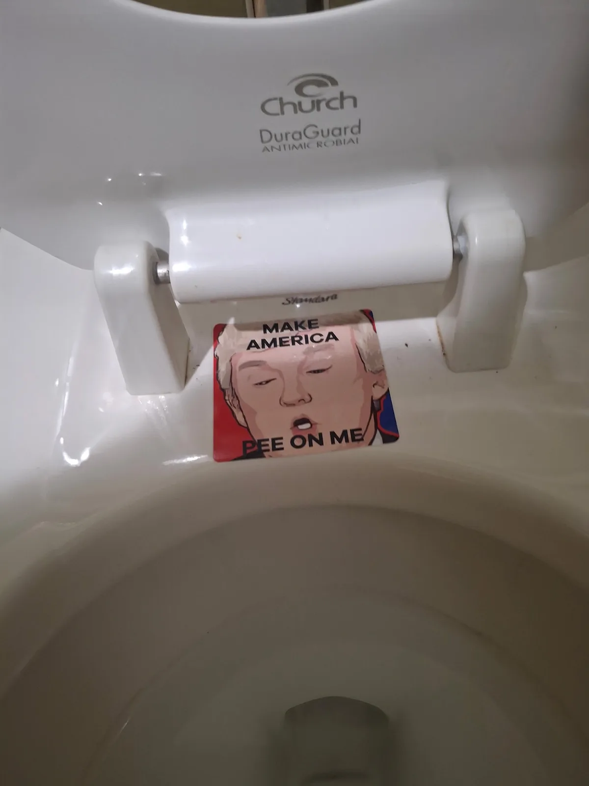 A close-up photo shows a white toilet with the lid open, revealing a sticker on the toilet seat hinge. The sticker features…
