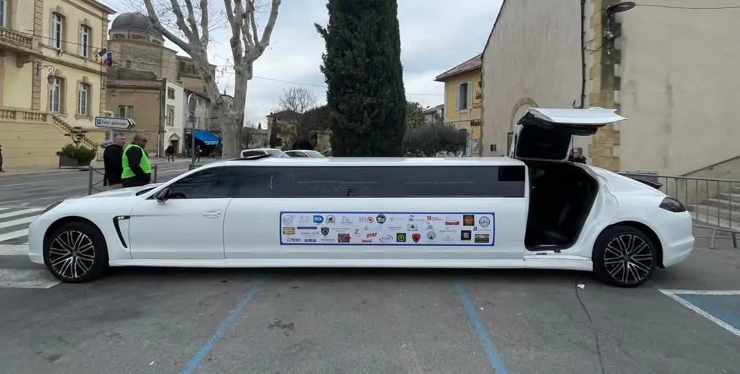 A white, elongated stretch Porsche is parked on a street with its rear door open, displaying a long row of sponsor logos…