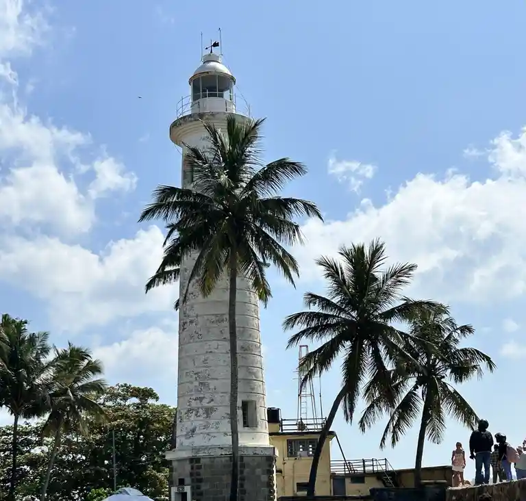 A tall, white, weathered lighthouse stands against a bright blue sky with scattered clouds, partially obscured by tall palm…
