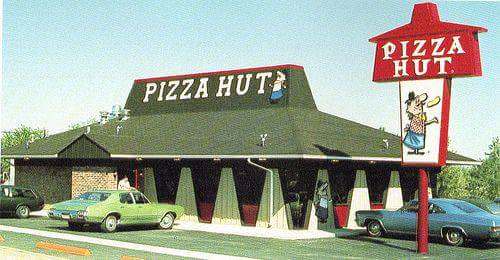 A vintage photo of a Pizza Hut restaurant from the 1960s or 70s, featuring its distinctive red and white sign with a cartoon…