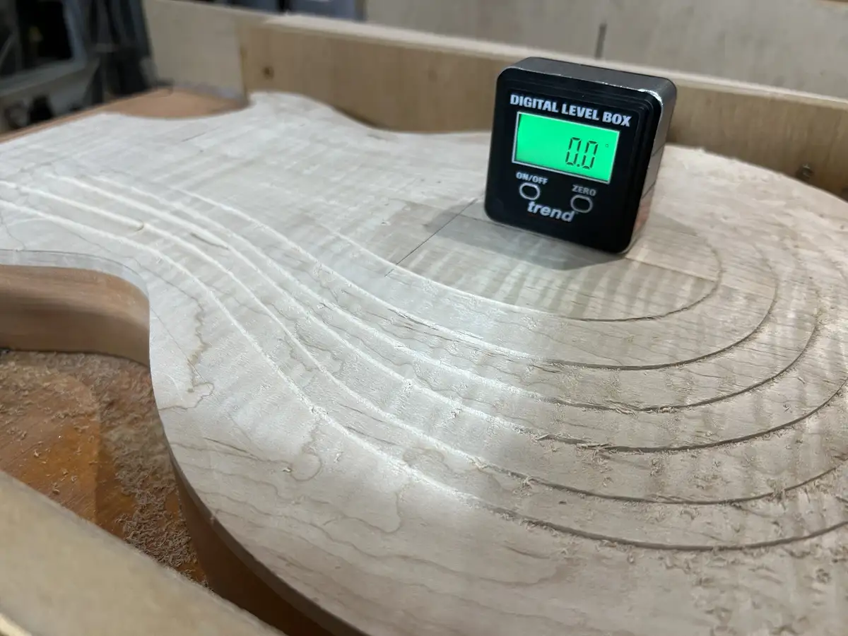 A black digital level box with a green screen displaying '0.0°' sits on a freshly sanded, unfinished guitar body made of…