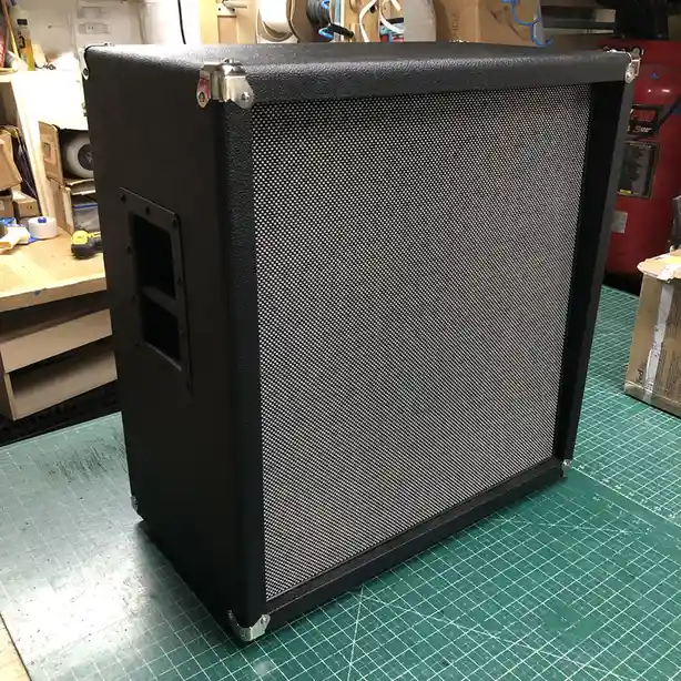 A black guitar amplifier cabinet with a perforated metal grille sits on a green cutting mat in a workshop. The text overlaid…