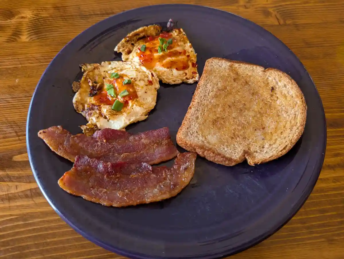 A plate of breakfast food including two fried eggs with red sauce and green onions, two strips of bacon, and one slice of…
