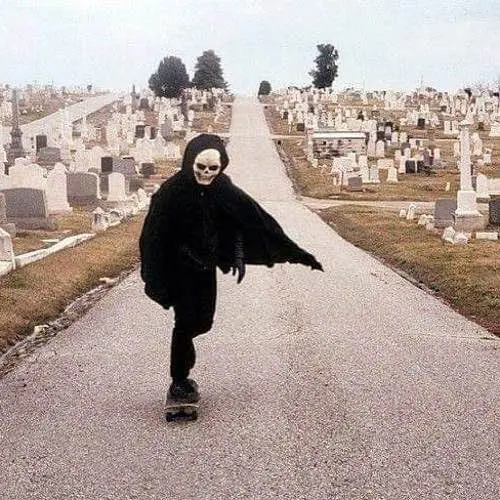 A person wearing a full skull mask and a black hooded cloak is skateboarding down a paved path through a cemetery filled…