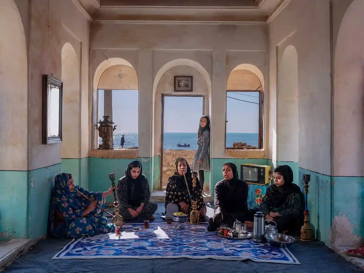 A group of women in traditional headscarves sit on a blue patterned rug inside a room with arched windows overlooking the…