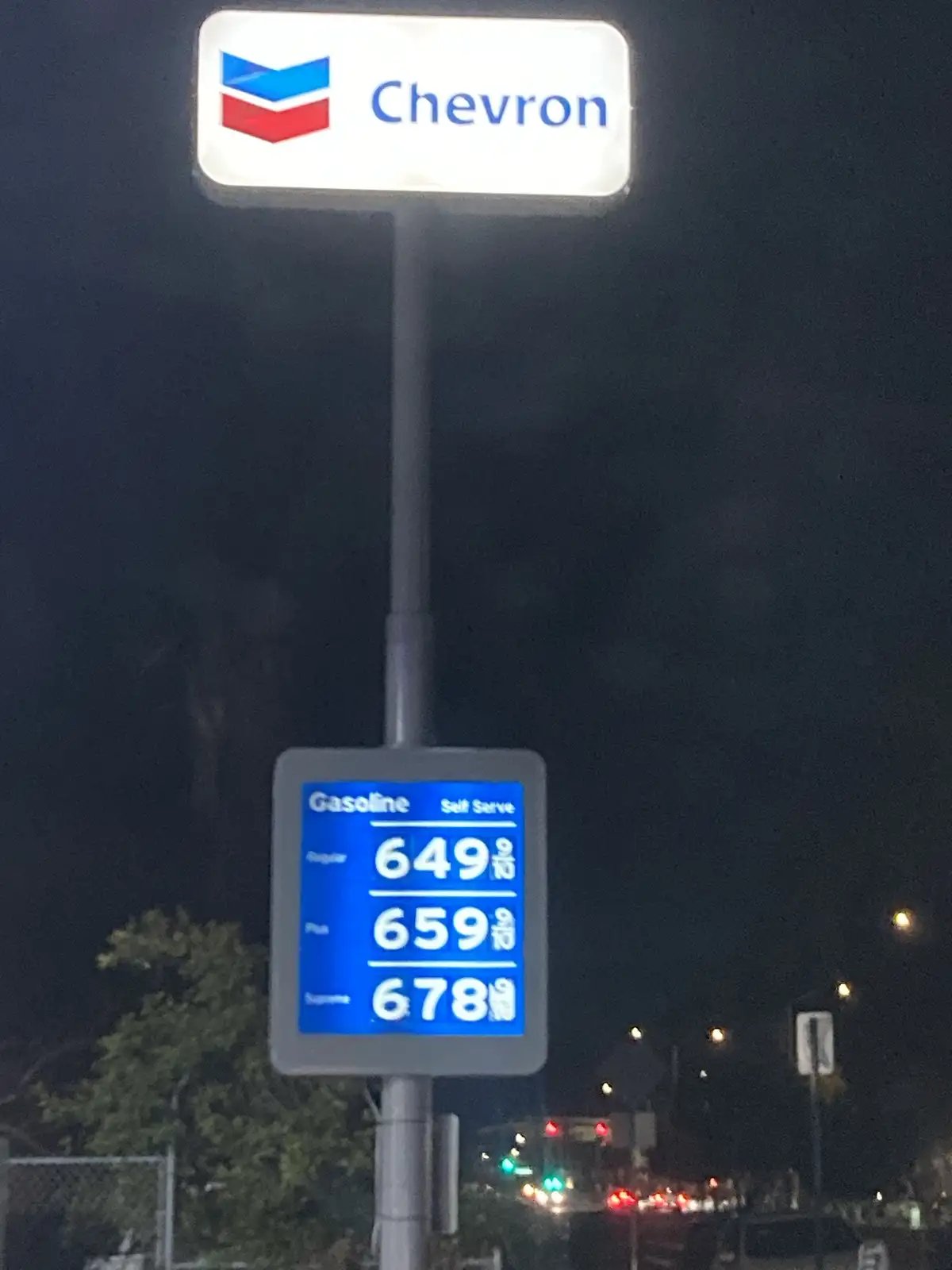 A nighttime photo of a Chevron gas station sign showing self-serve gasoline prices: Regular at 649.9, Plus at 659.9, and…