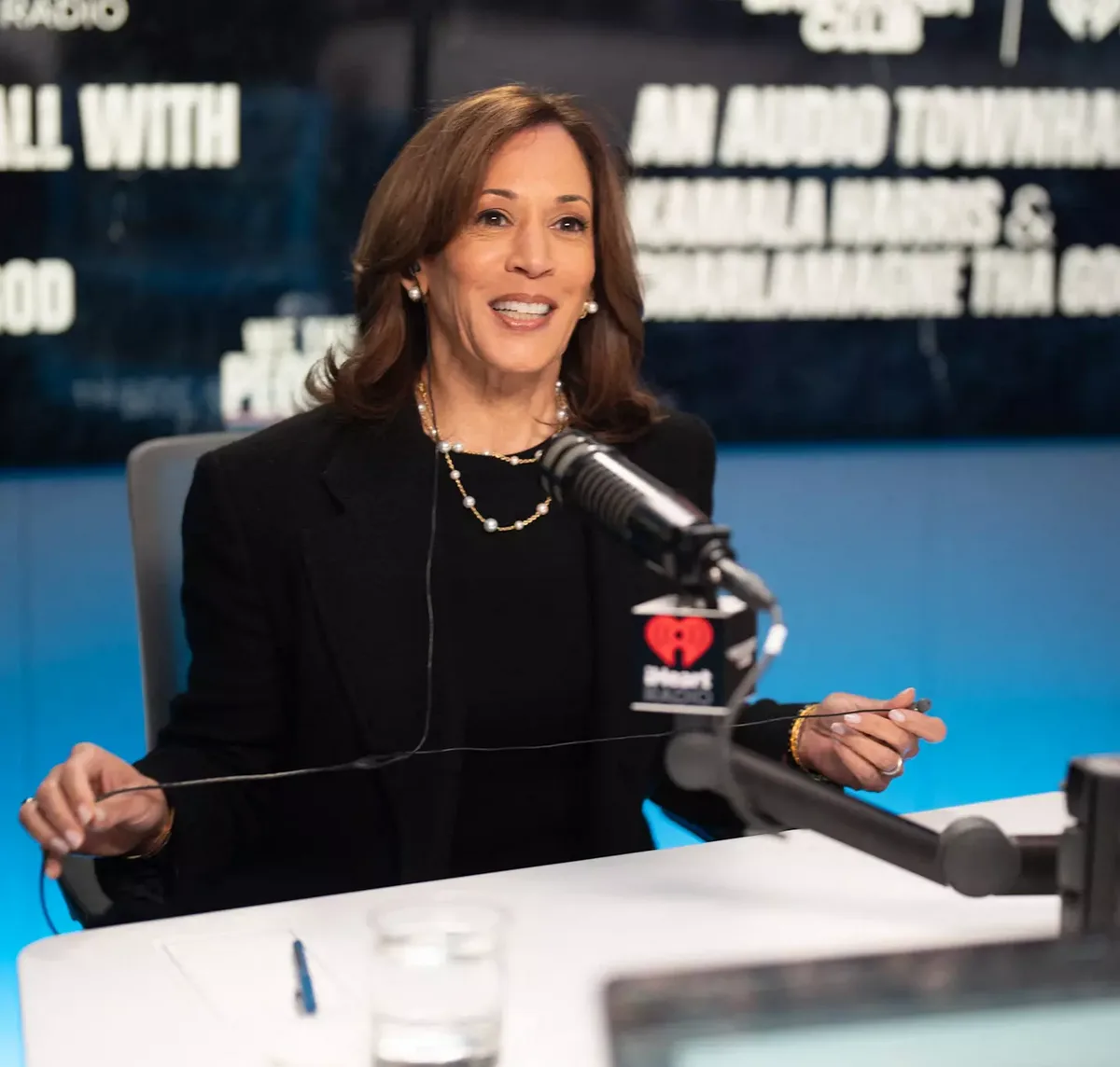 kamala harris is seated at a white desk in front of a microphone with an iheart radio logo, smiling while holding a thin…