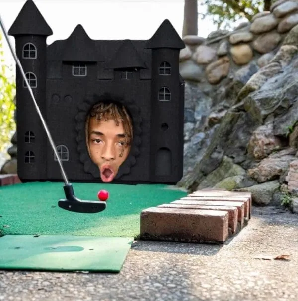 A miniature golf scene features a black castle-shaped hole with a cutout face of a man with an open-mouthed expression. A…