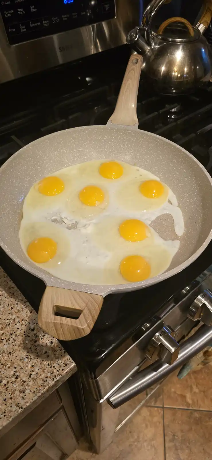 A frying pan on a stovetop contains seven raw eggs, their yolks intact and surrounded by cooking whites, with a stainless…