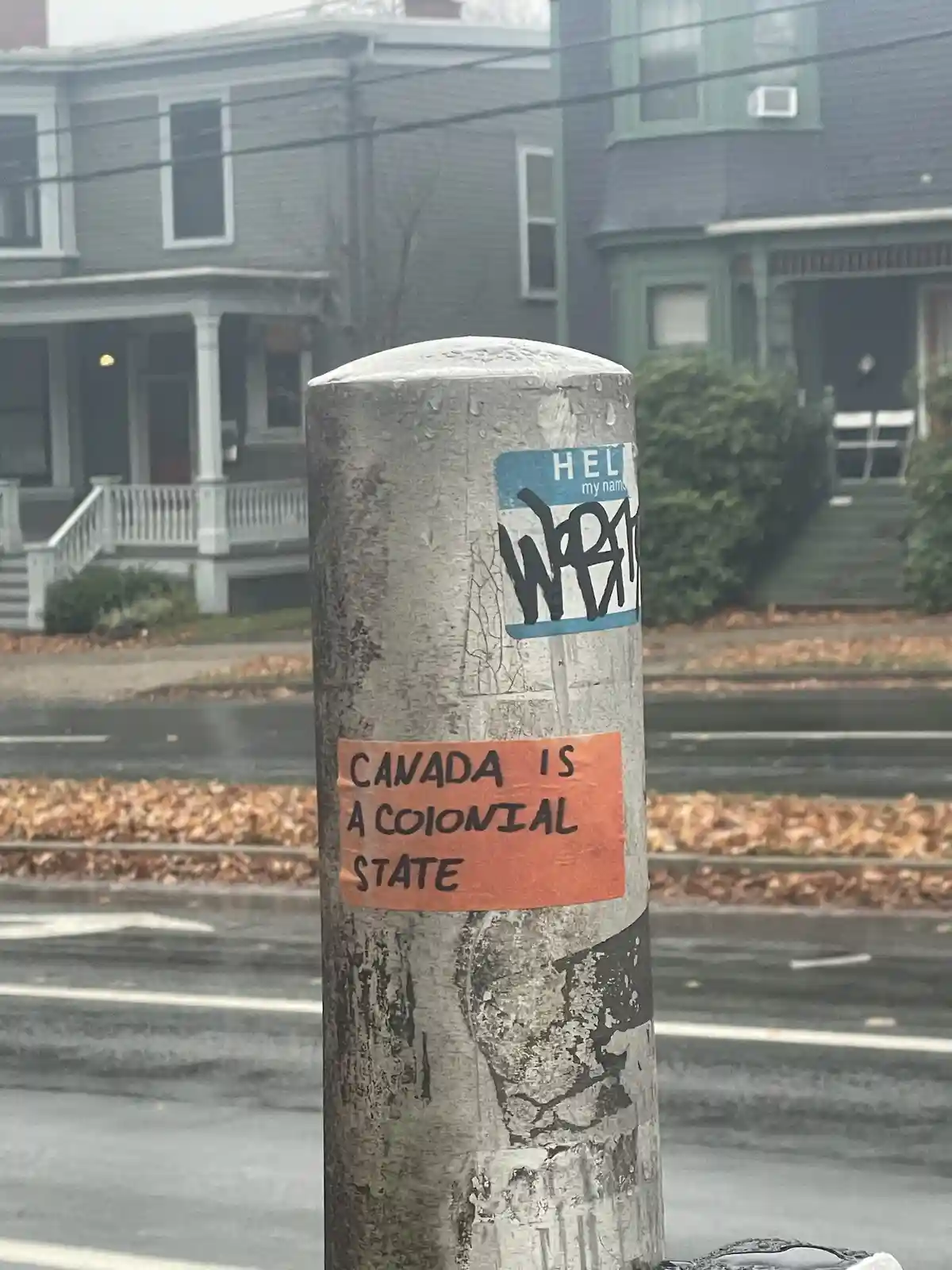 A weathered utility pole covered in stickers and graffiti stands on a wet street with houses in the background. A prominent…