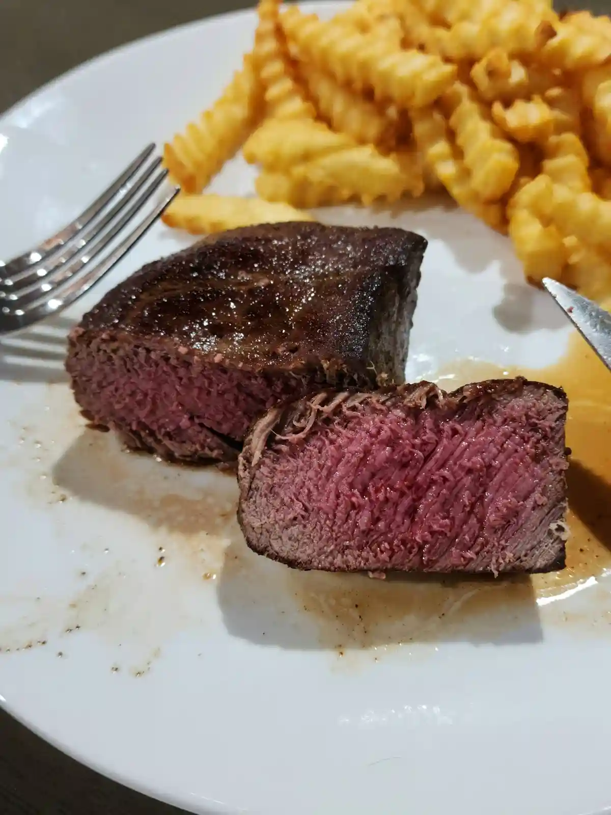 A close-up photo shows a sliced, rare steak on a white plate with crinkle-cut fries, a fork on the left, and a knife on the…