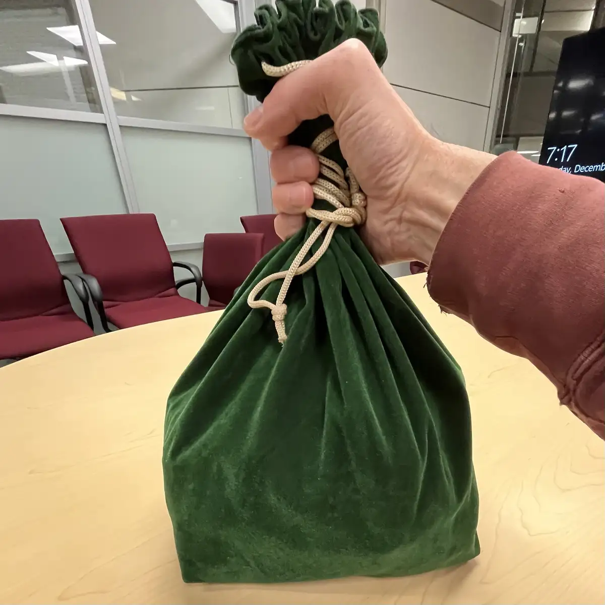 A hand holds a green velvet drawstring bag on a wooden table in an office setting with red chairs and glass walls; a digital…