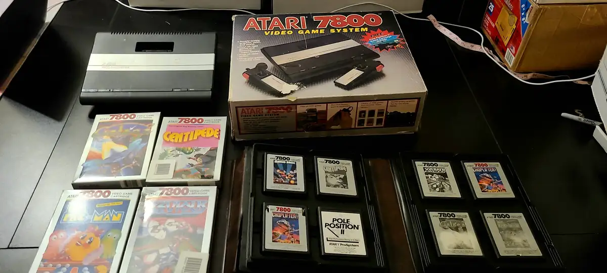 An Atari 7800 video game console sits next to its original box and a collection of game cartridges and boxes, including…