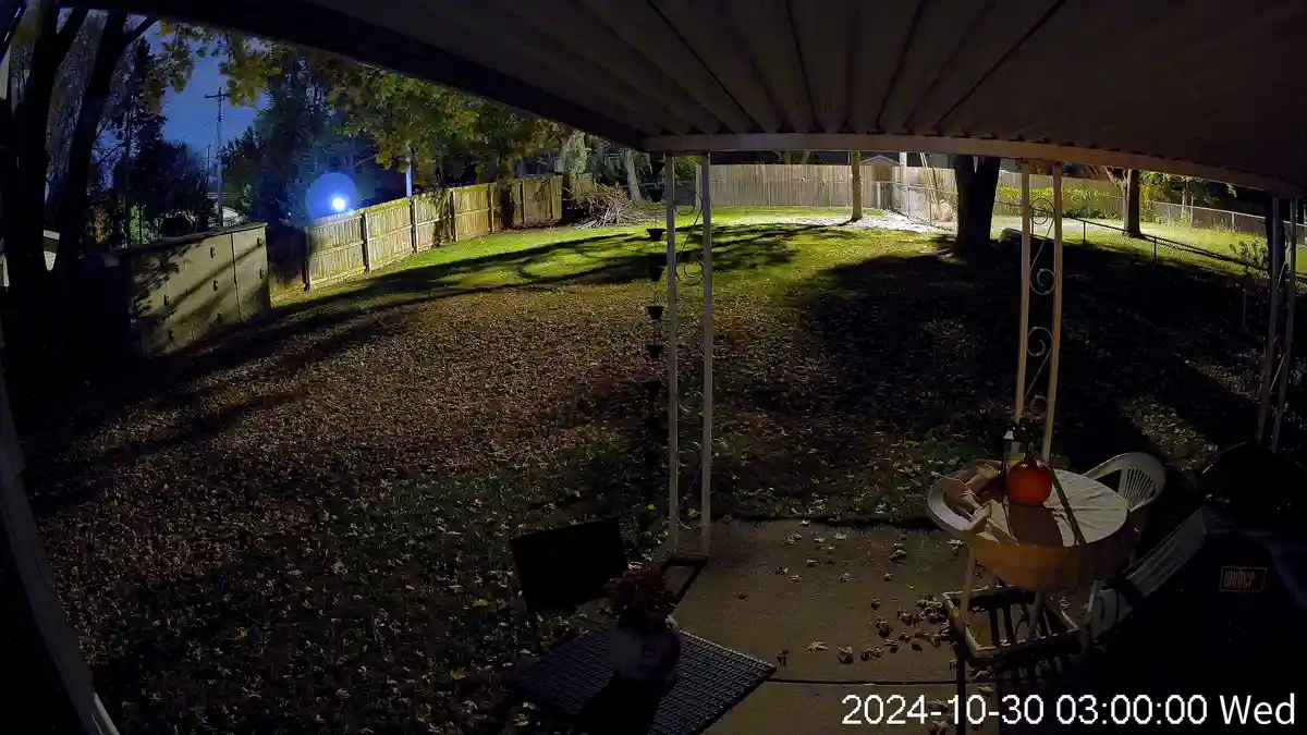 A nighttime backyard security camera view shows a lit patio with tables, chairs, and a pumpkin under a pergola, surrounded…