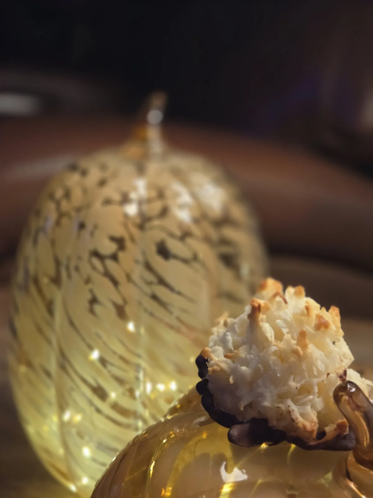 A close-up photo shows a coconut macaroon resting on a golden, ornate glass pumpkin-shaped container, with another similar…