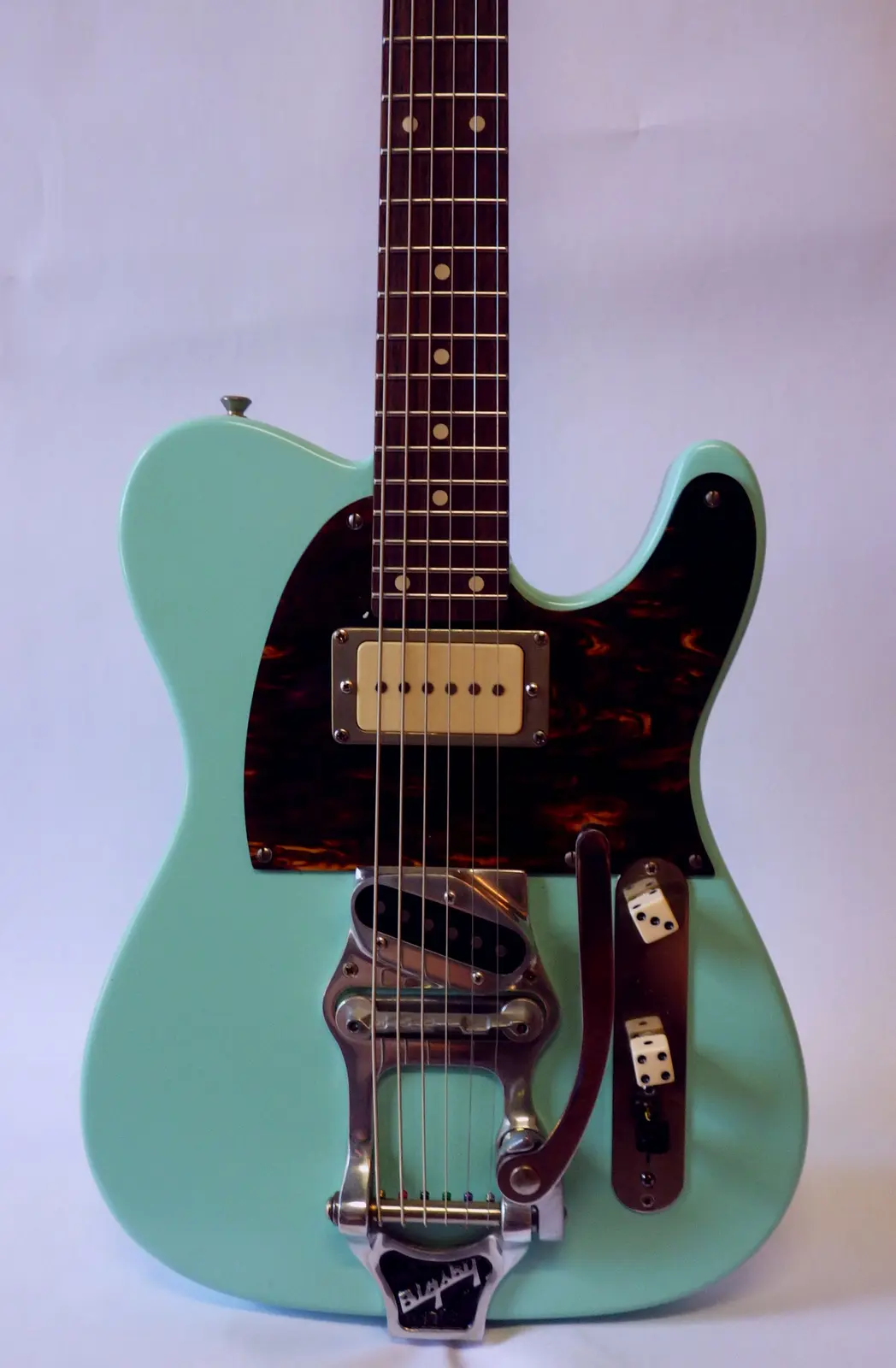 A mint green electric guitar with a tortoiseshell pickguard and a Bigsby vibrato tailpiece, modified with two dice mounted…