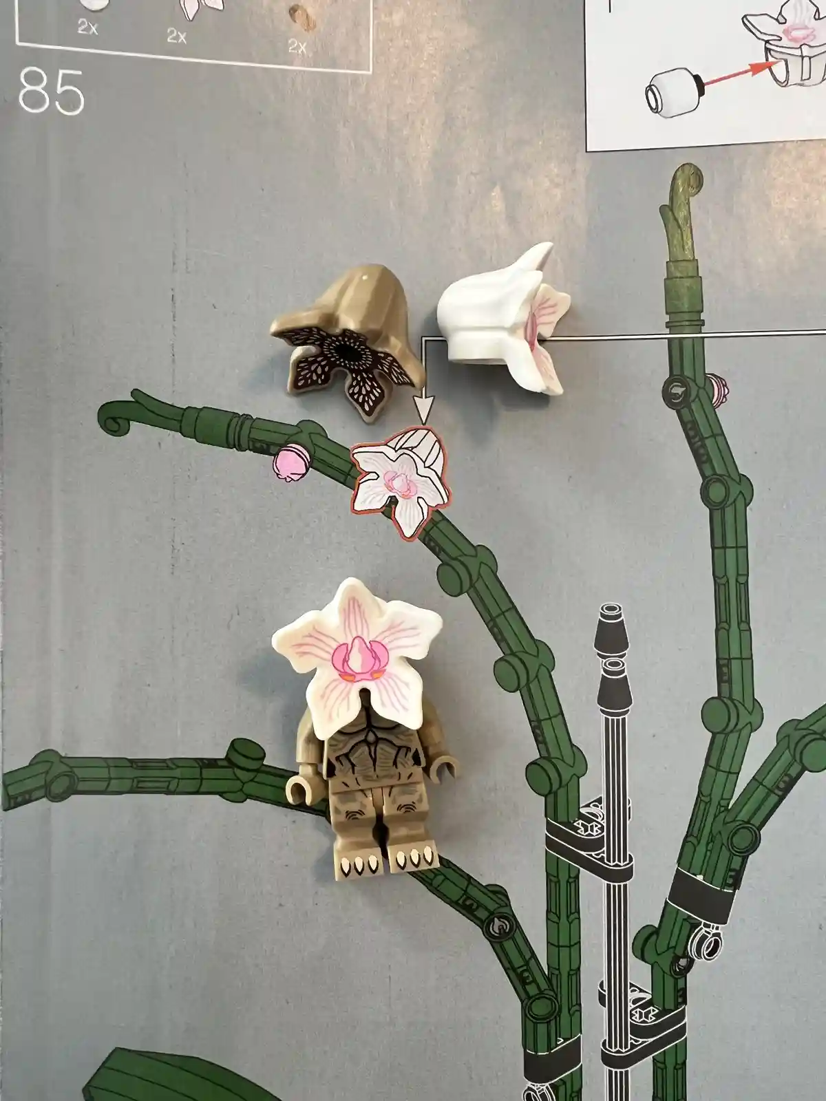 A close-up of a LEGO instruction manual page showing a tan minifigure with a large white orchid flower piece attached to its…