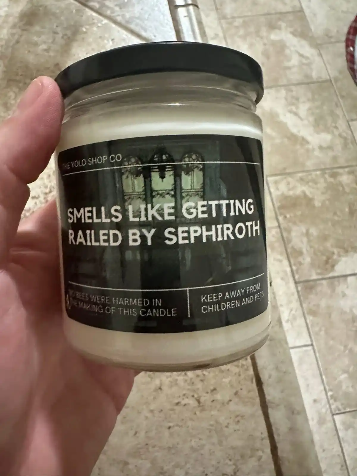 A hand holds a glass jar candle with a black label that reads 'SMELLS LIKE GETTING RAILED BY SEPHIROTH' in large white text…