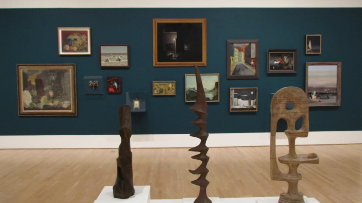 A museum gallery with a dark teal wall displaying numerous framed paintings of varying sizes and styles. In the foreground…