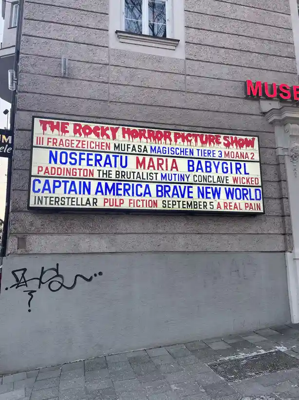 A theater marquee displays a surreal movie lineup including 'The Rocky Horror Picture Show' at the top, followed by a…