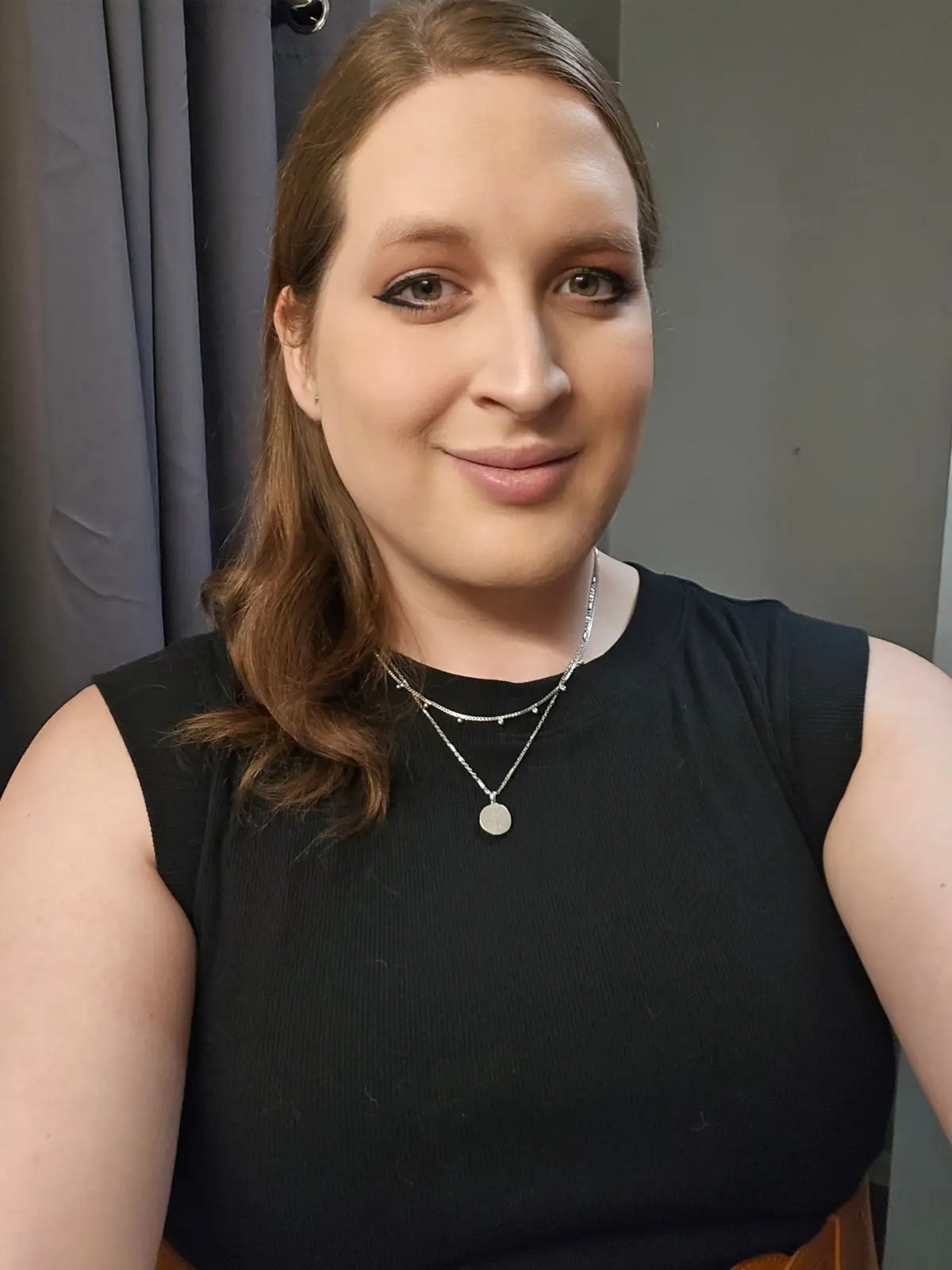 A woman with brown hair and makeup smiles at the camera while wearing a black sleeveless top and layered silver necklaces…